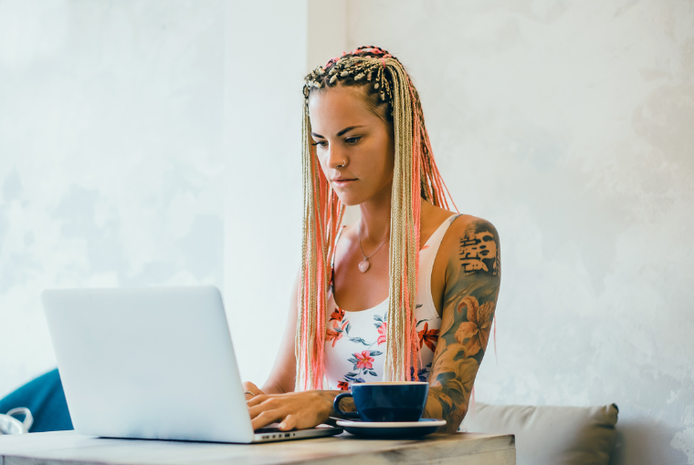 Tattooed woman working on a laptop from a home office with a big cup of coffee