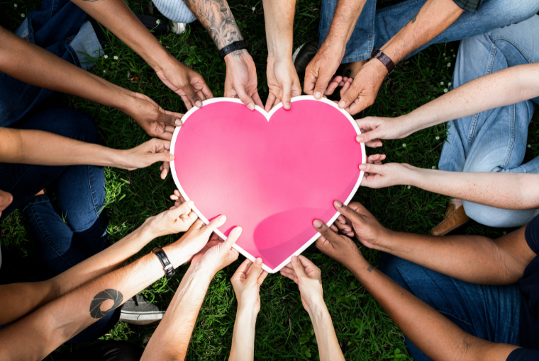 A large pink paper heart is in the centre of a lawn and help by multiple people's hands