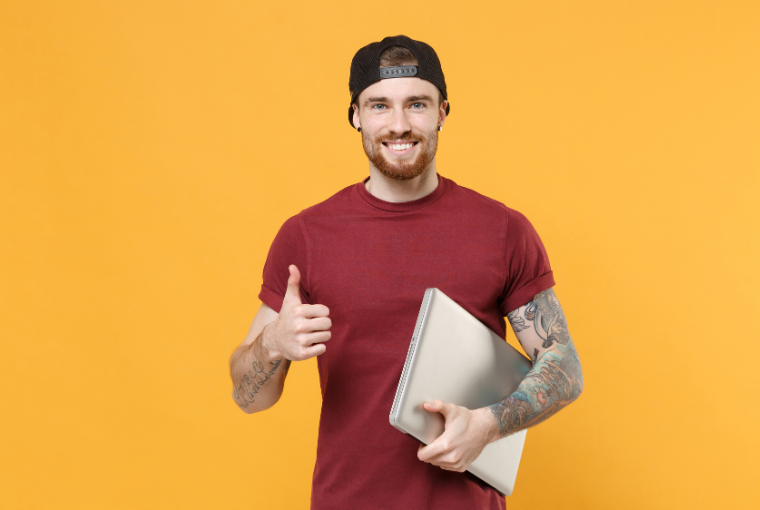 A young man in a baseball cap clutches a laptop under his arm and gives a thumbs up to the camera