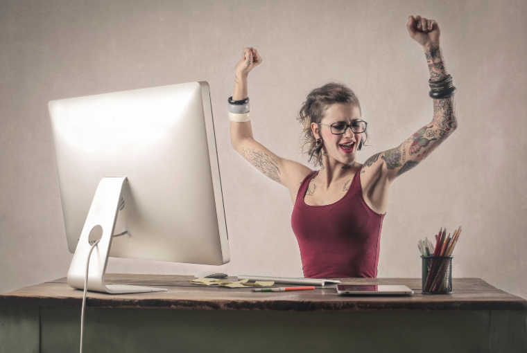 Tattooed woman sitting at a desk with a computer smiles and raises her arms 