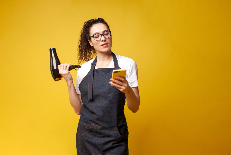 A hairdresser stands against a bright yellow background with a hair dryer in one had and a mobile phone in the other 