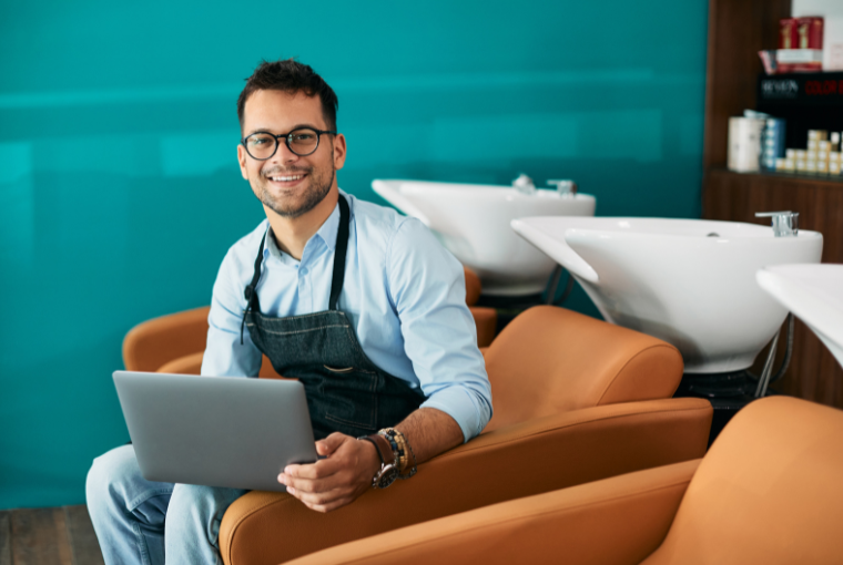 A male stylist sitting in front of a row of sinks holding a laptop and smiling