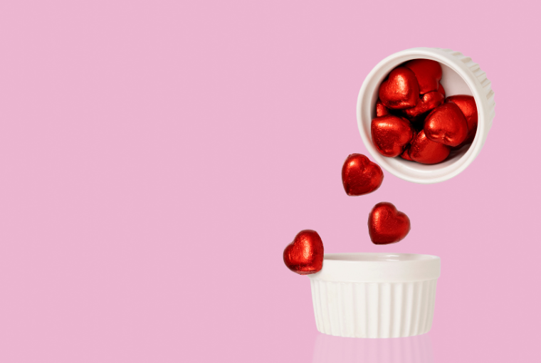 A bowl of red heart shaped candies fall from a bowl on a soft pink background