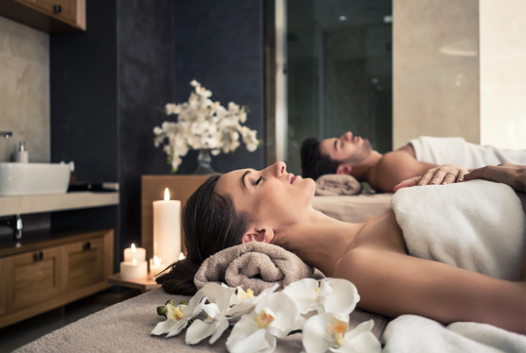 A relaxing couple lie down at a spa with white flowers and candles, wrapped in fluffy white towels