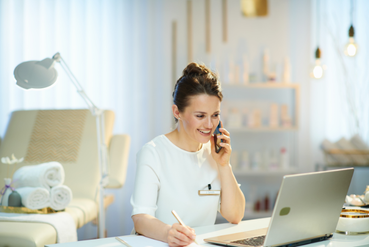 Happy spa employee chats to a customer on the phone as she uses spa management software to take a booking on her laptop